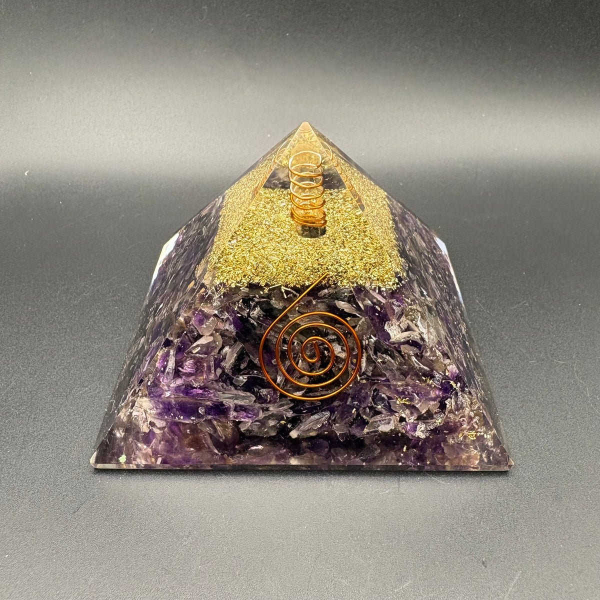 A resin triangular pyramid tower made of layered materials, with a gold glitter top section, Amethyst base, and a copper spiral design on the front, photographed on a dark gray background.