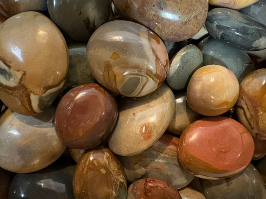 A close-up pile of polished Mookite stones ; earthy tones, including tan, brown, gray, muted red, and cream, with smooth rounded surfaces and visible natural banding.