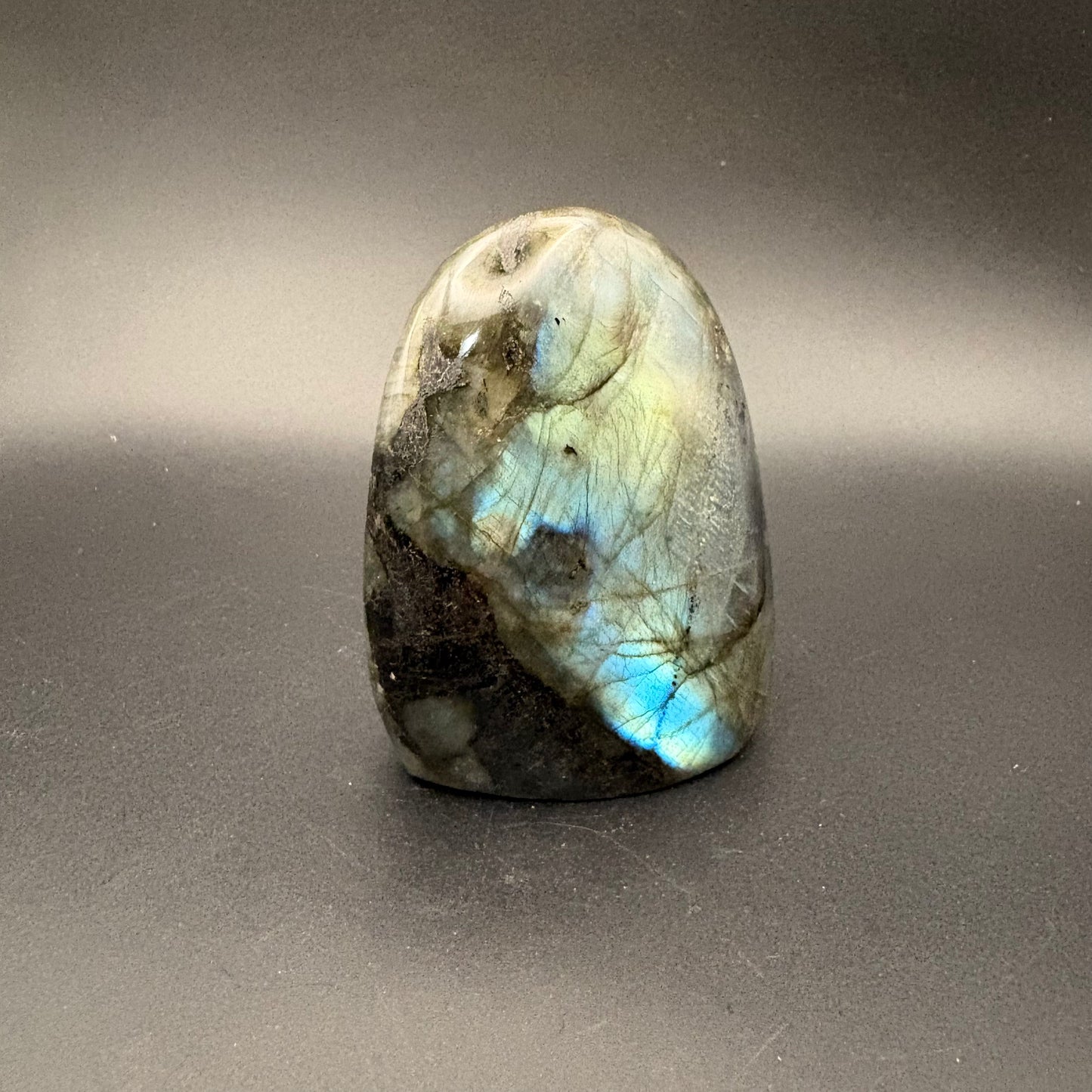 A polished labradorite crystal stone with natural inclusions and a vibrant blue iridescent flash, standing upright on a dark gray background.