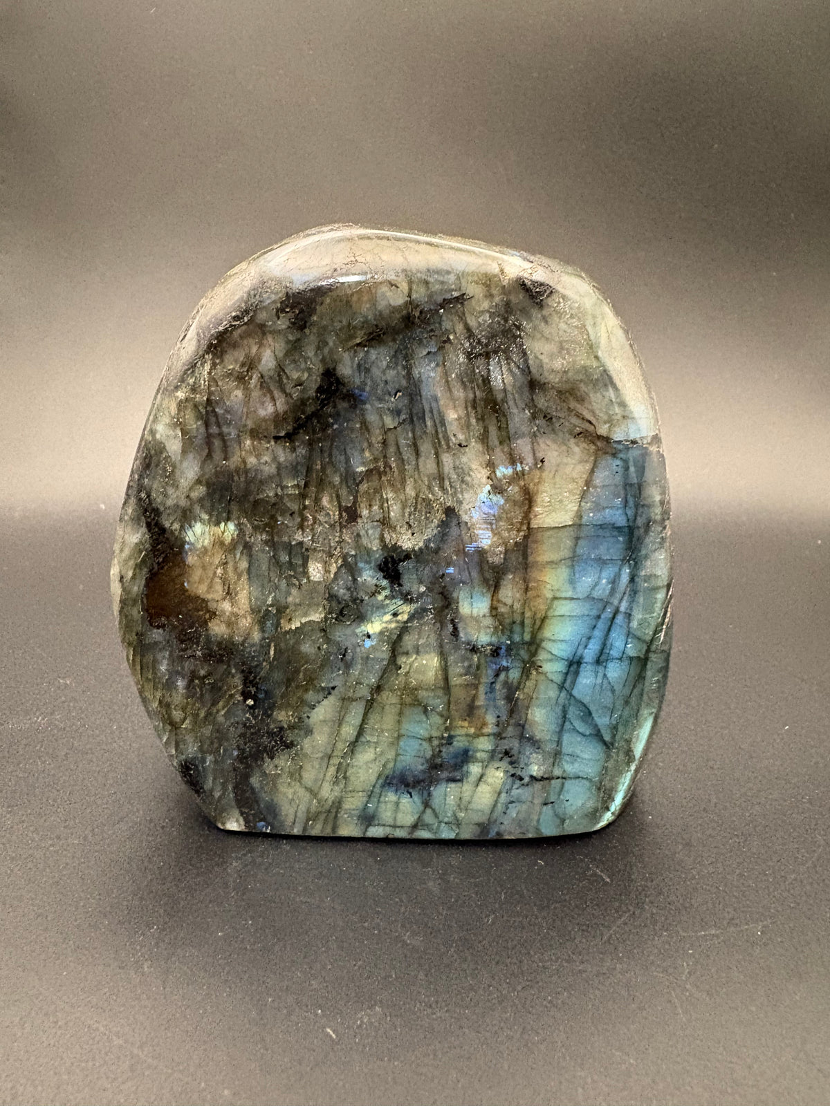 A polished natural labradorite stone with iridescent blue, green, and gold flashes, set against a dark gray background.