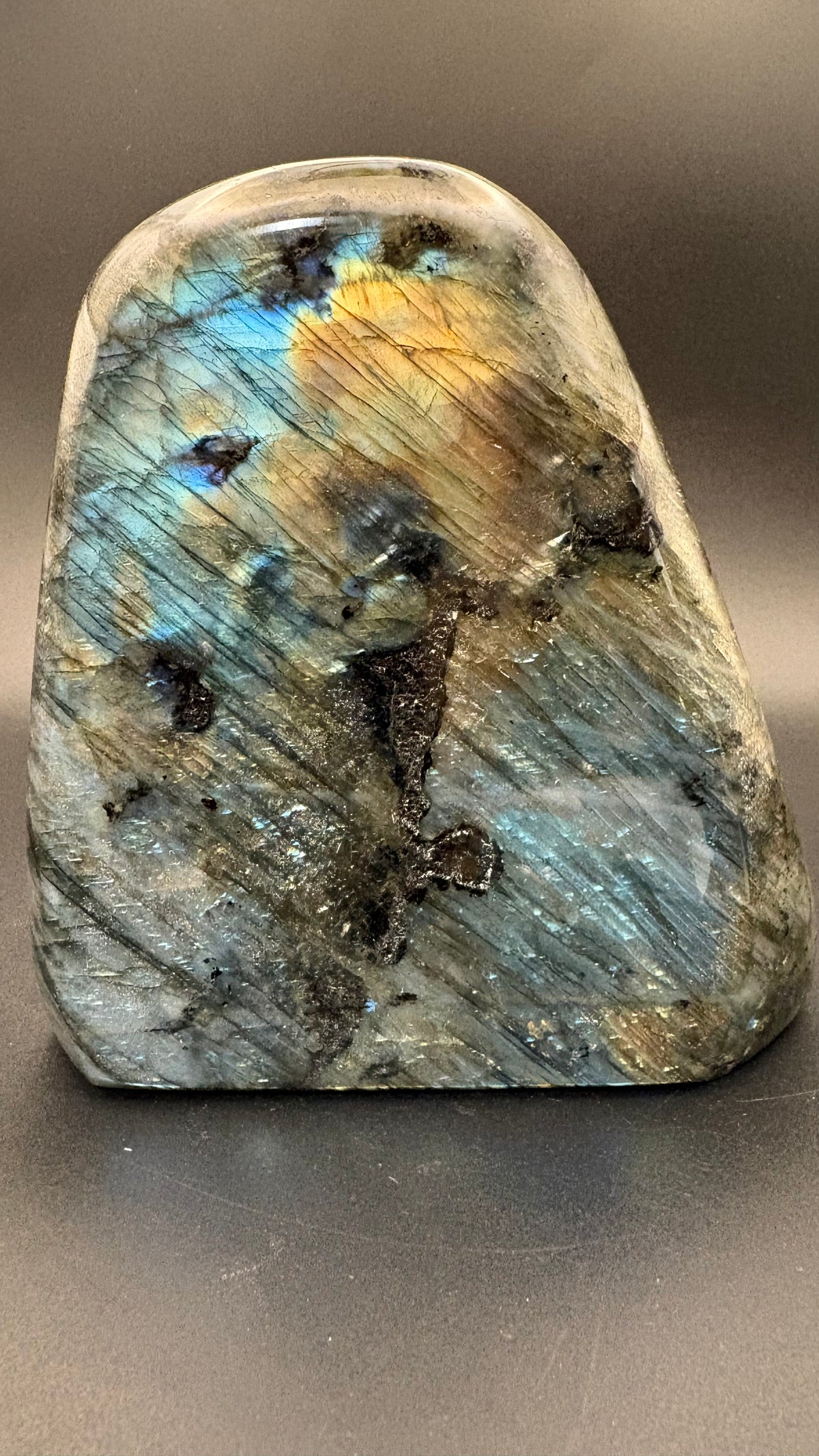 A polished natural labradorite stone with iridescent blue, gold, and gray flashes, photographed on a dark gray surface.