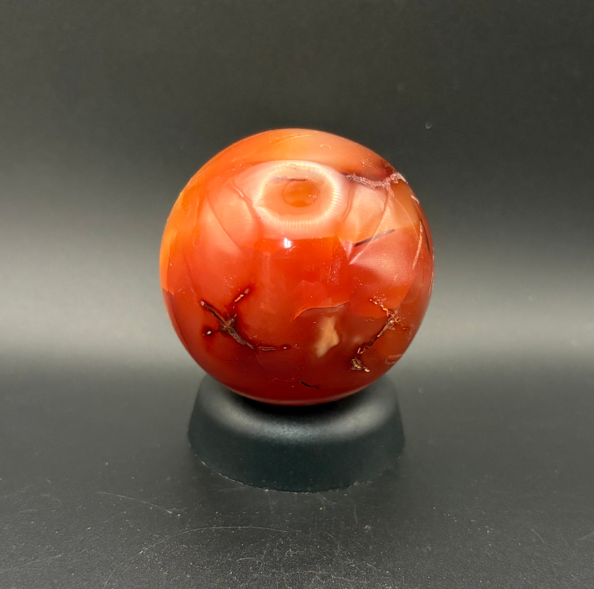 A polished Carnelian spherenwith natural inclusions, displayed on a small black stand against a dark gray background.