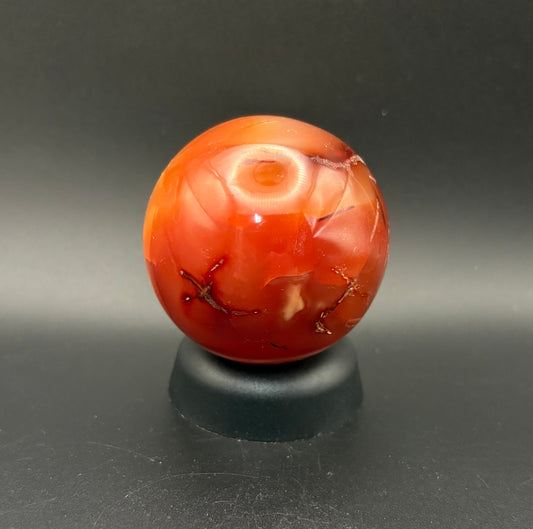 A polished Carnelian spherenwith natural inclusions, displayed on a small black stand against a dark gray background.