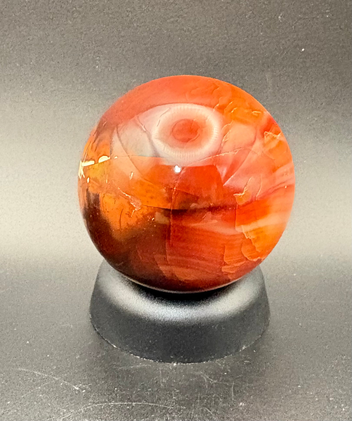 A polished Carnelian spherenwith natural inclusions, displayed on a small black stand against a dark gray background.