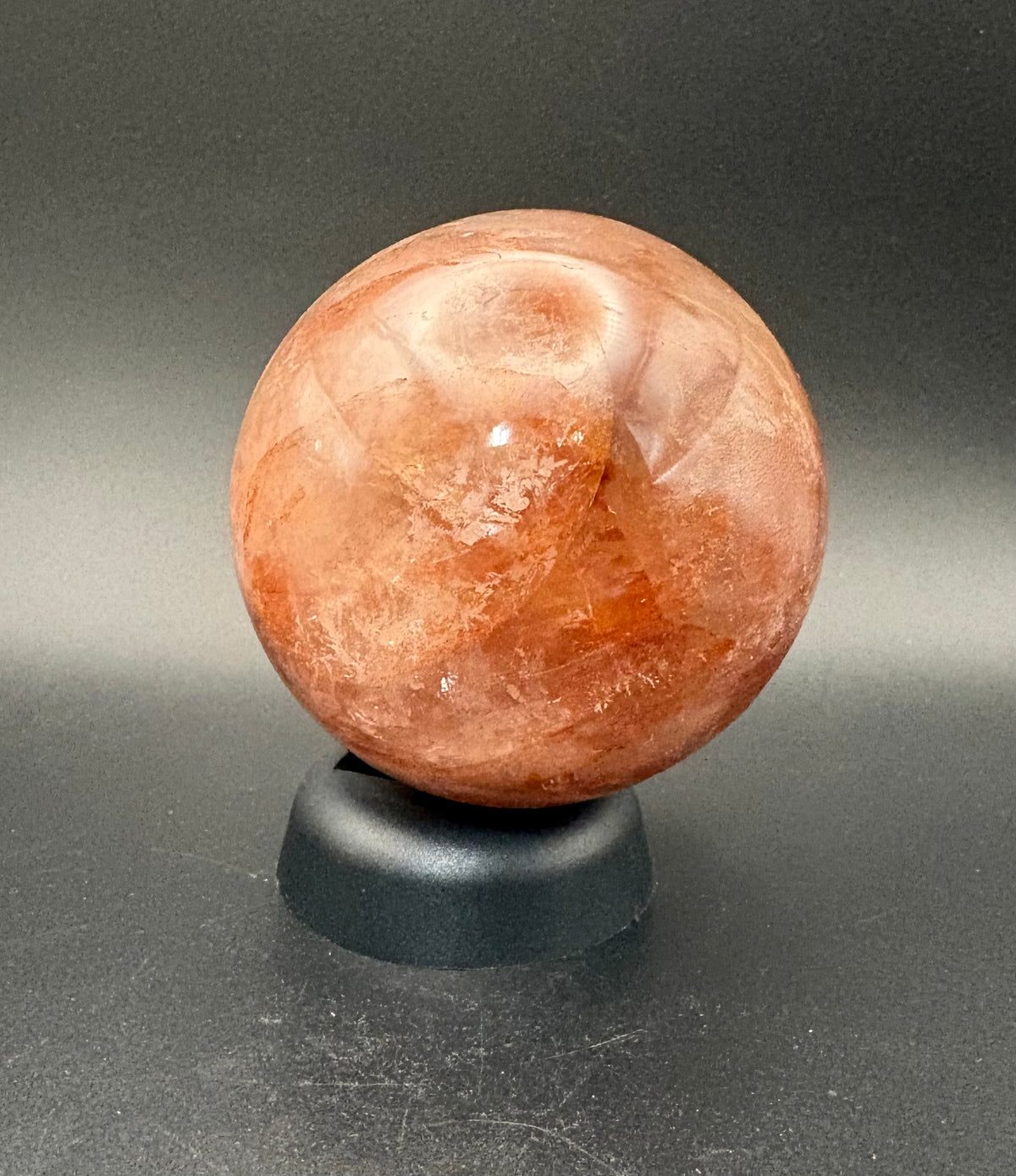 A polished fire  quartz crystal sphere resting on a small black display stand against a dark gray gradient background.