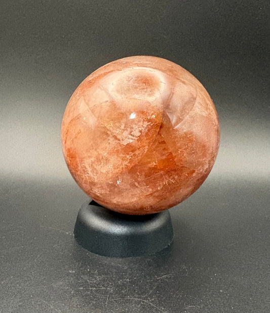 A polished fire  quartz crystal sphere resting on a small black display stand against a dark gray gradient background.