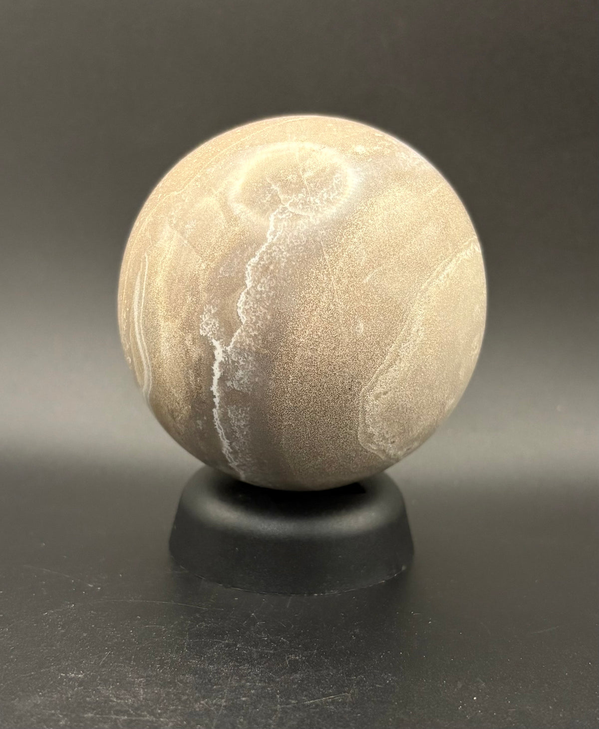 A polished beige Moonstone sphere with natural white veining, displayed on a small black stand against a dark gray background.