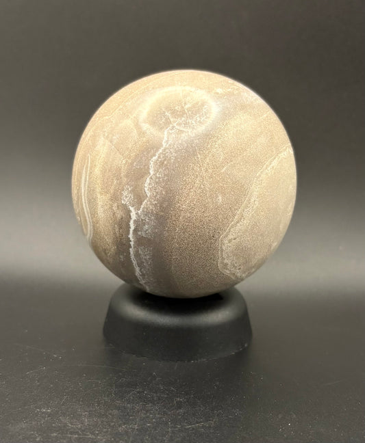 A polished beige Moonstone sphere with natural white veining, displayed on a small black stand against a dark gray background.