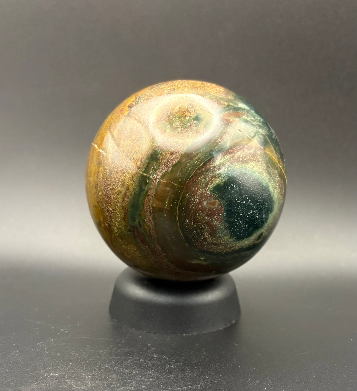 A polished Mookite stone sphere with swirling patterns of brown, gold, green, and dark blue, displayed on a small black stand against a dark gray gradient background.