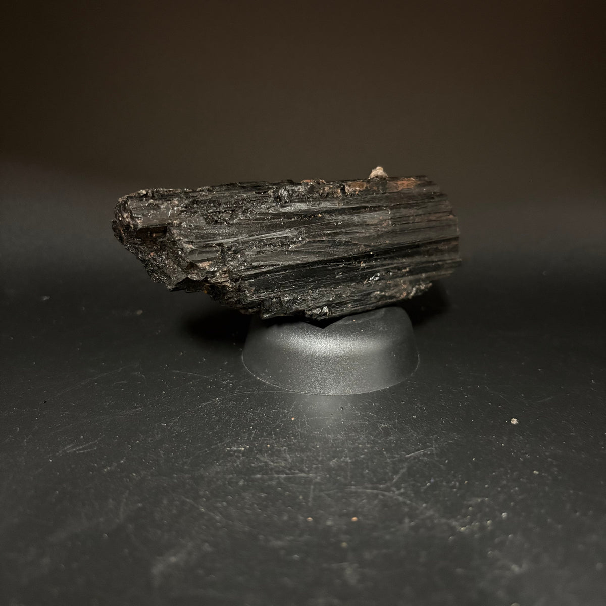 A dark, tourmaline specimen or  displayed on a small round black stand against a dark studio background.