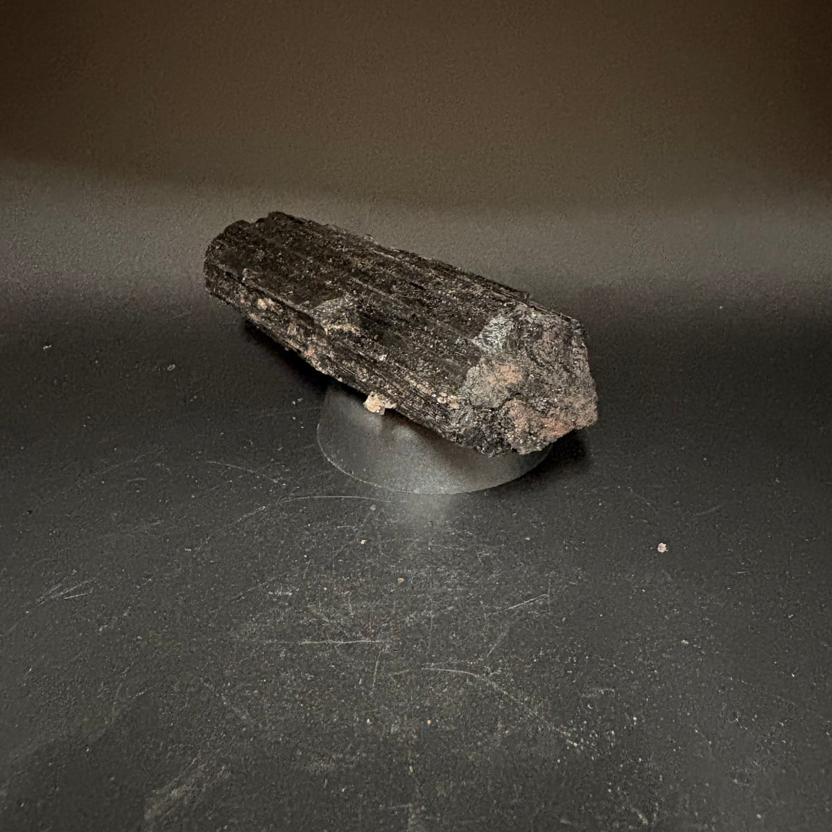 A rough, tourmaline crystal specimen displayed on a small clear stand against a dark gray studio background.