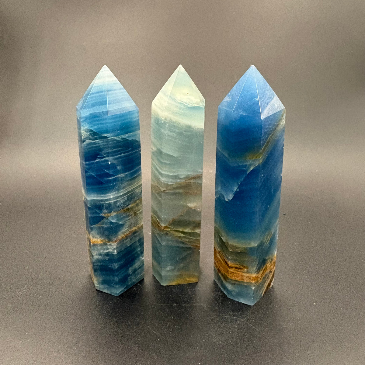 Three polished blue stone crystal towers with natural banding and gold veining, standing upright on a dark gray surface against a neutral gray background.