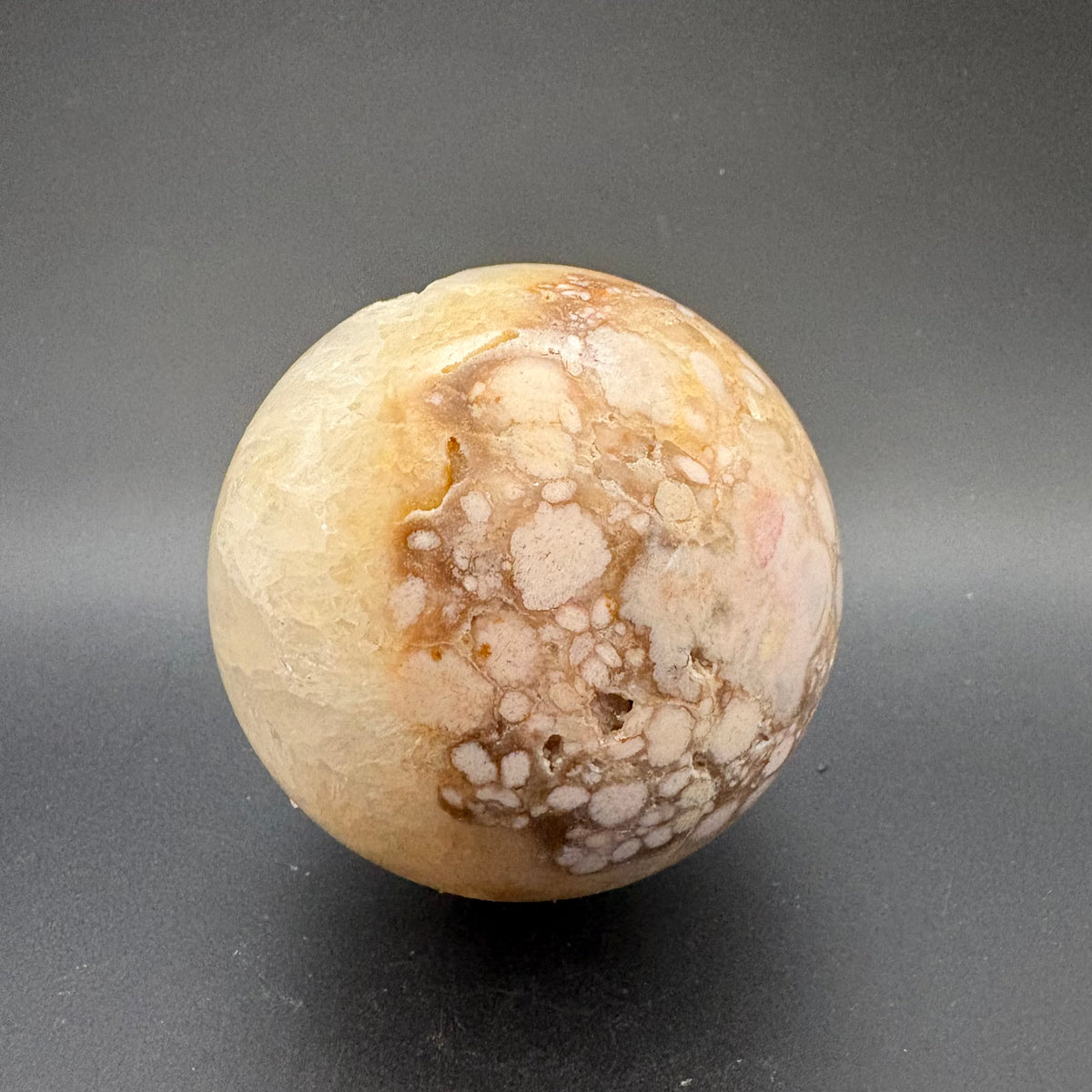 A polished spherical flower agate stone with a marbled pattern of cream, beige, and brown, standing on a dark gray surface.