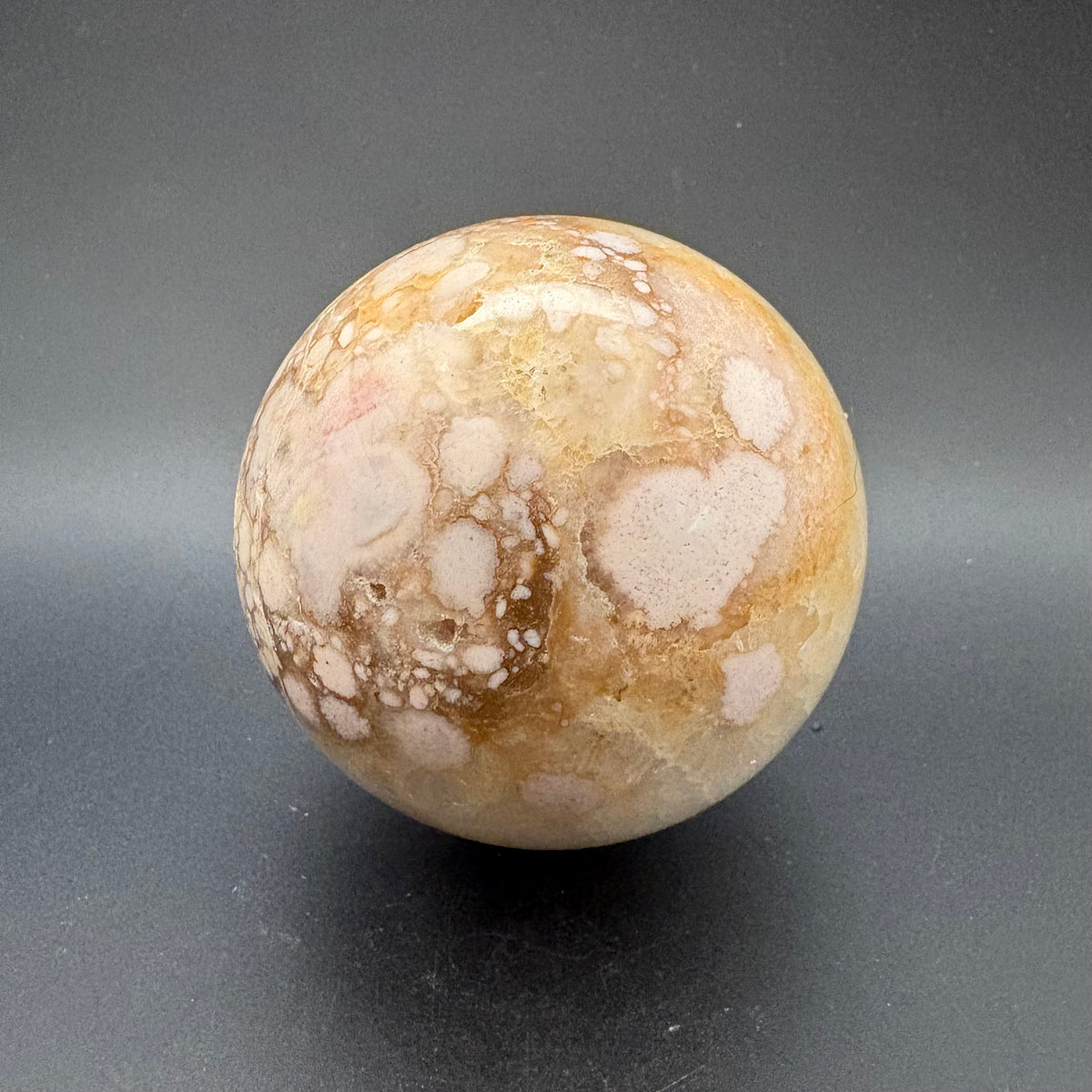 A polished spherical flower agate stone with a marbled pattern of cream, beige, and brown, standing on a dark gray surface.