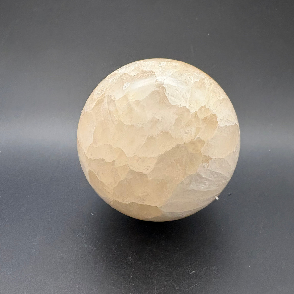 A polished spherical flower agate stone with a marbled pattern of cream, beige, and brown, standing on a dark gray surface.