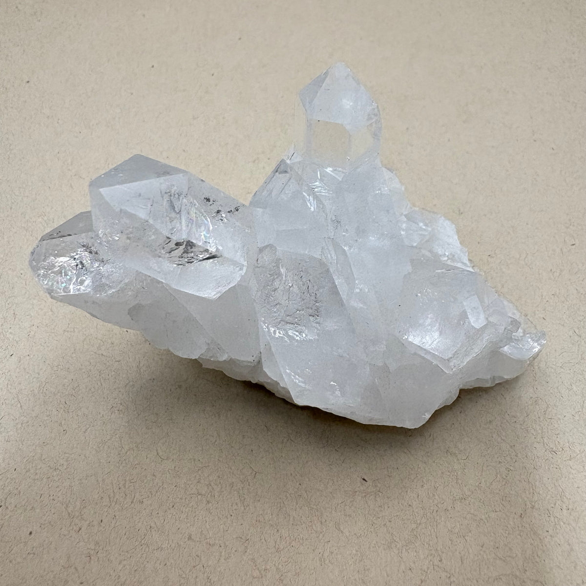 A natural clear quartz crystal cluster with multiple sharp points, displayed on a neutral beige textured surface.
