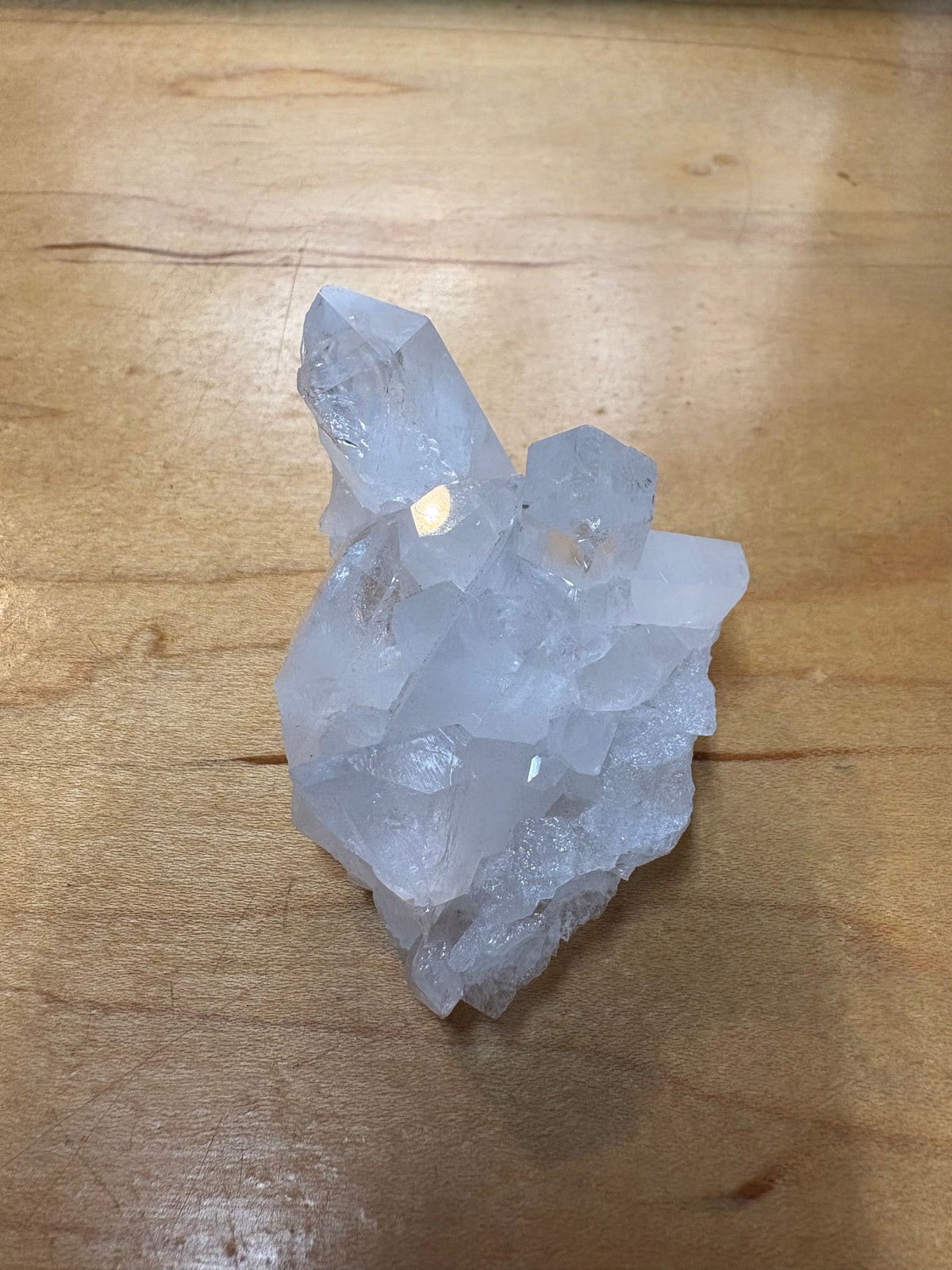A natural clear quartz crystal cluster with rough, unpolished facets, resting on a light brown wooden surface.