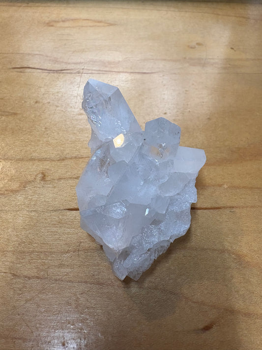 A natural clear quartz crystal cluster with rough, unpolished facets, resting on a light brown wooden surface.