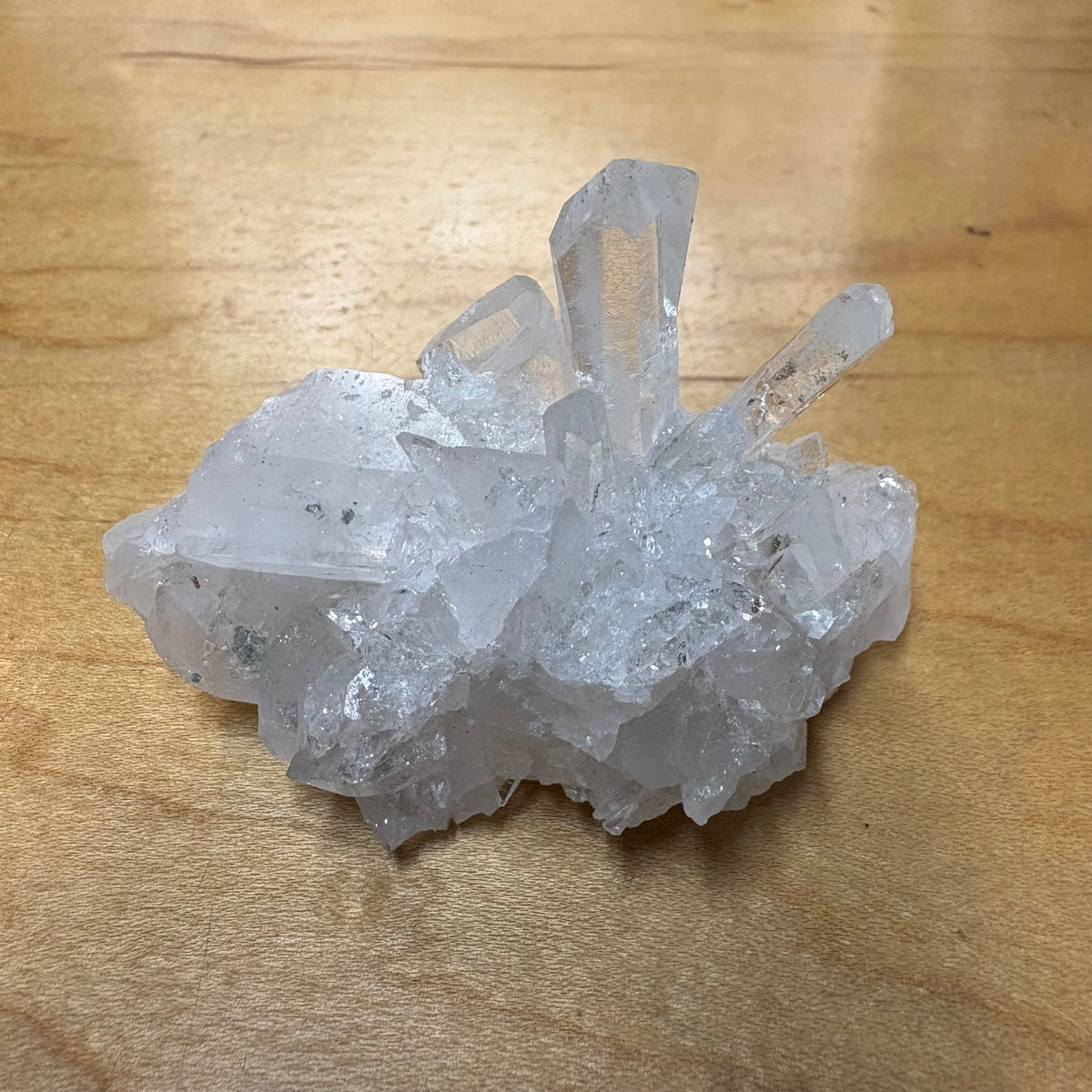 A natural clear quartz crystal cluster with multiple pointed crystal points, resting on a light brown wooden surface.