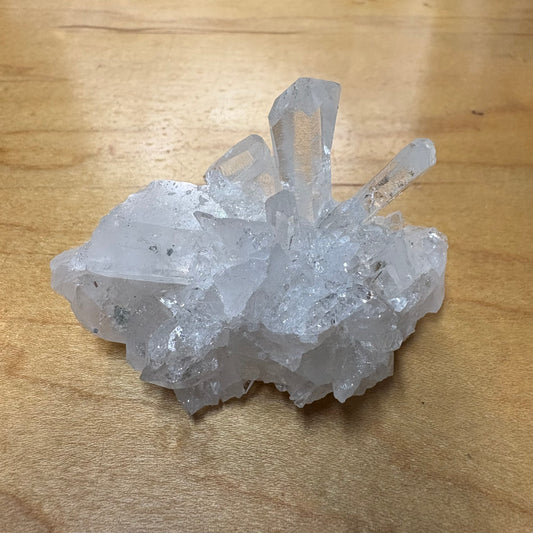 A natural clear quartz crystal cluster with multiple pointed crystal points, resting on a light brown wooden surface.