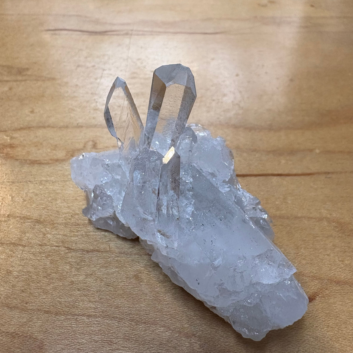 A natural clear quartz crystal cluster with multiple pointed crystal points, resting on a light brown wooden surface.