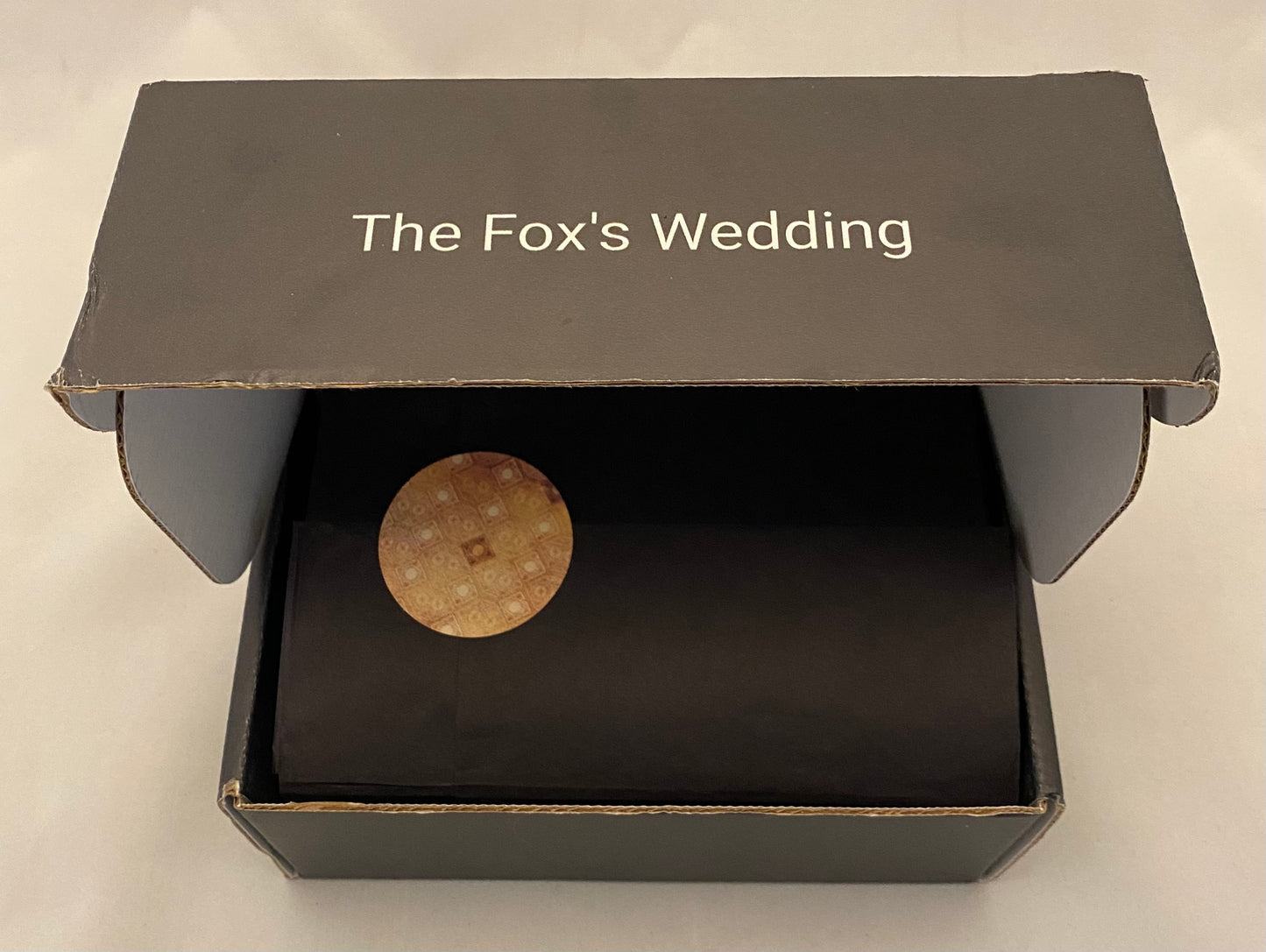 Black box with 'The Fox's Wedding' text and a circular object inside on a light gray background