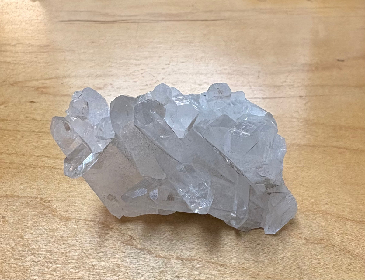 A raw, unpolished cluster of clear quartz crystals resting on a light-colored wooden surface.