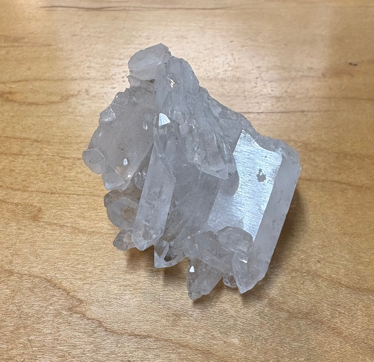 A raw, unpolished cluster of clear quartz crystals on a light brown wooden surface.