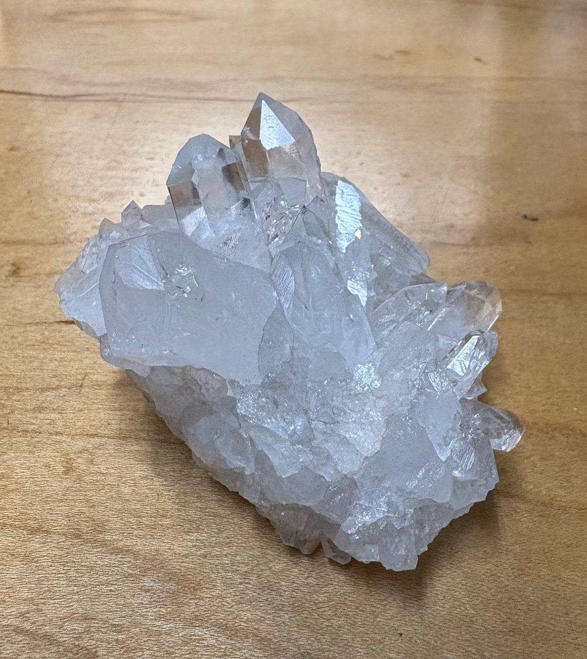A natural clear quartz crystal cluster with sharp geometric points, resting on a light brown wooden surface.