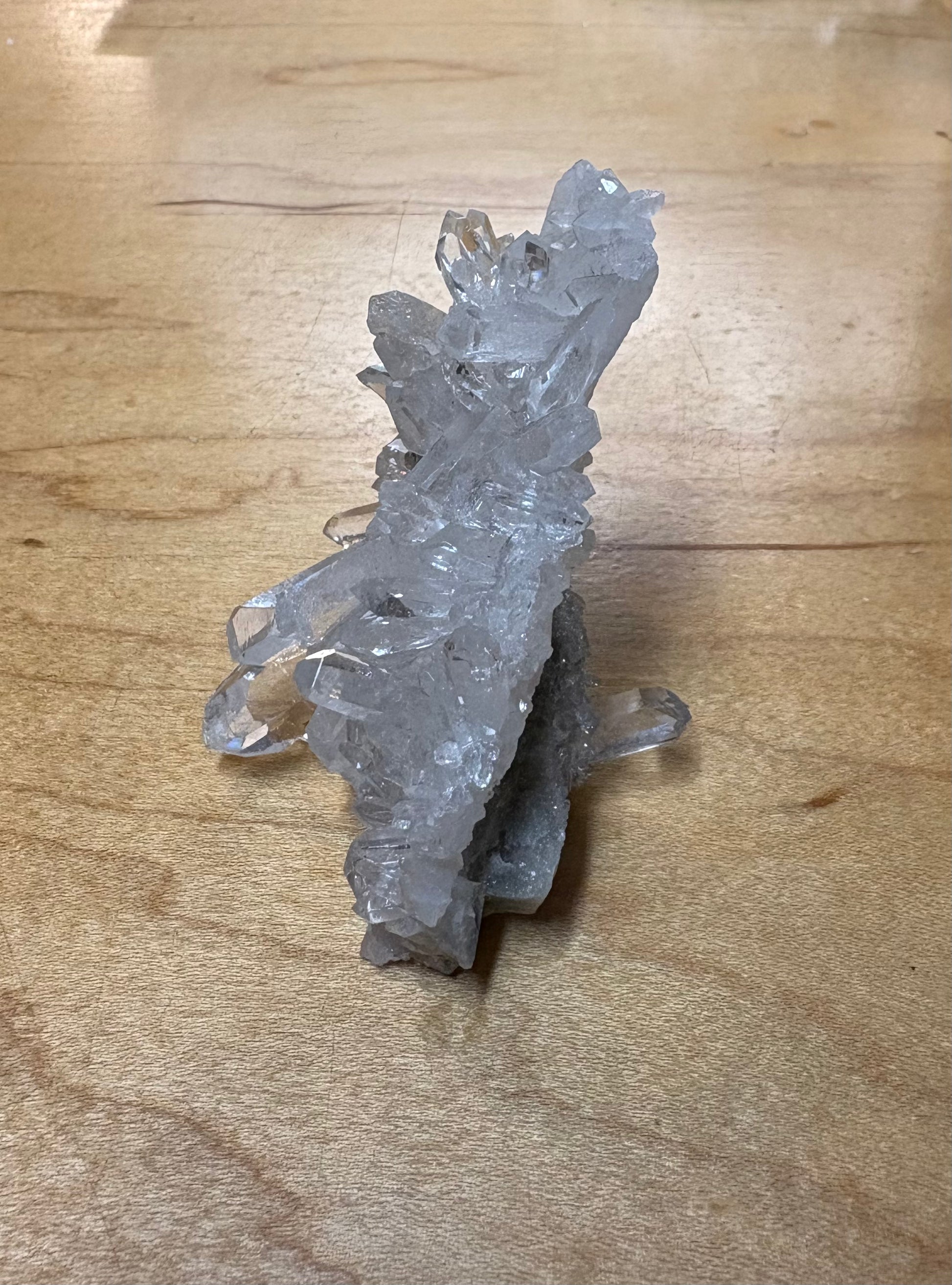 A raw crystal cluster, likely quartz, with clear and slightly grayish-blue crystalline structures, resting on a light brown wooden surface.