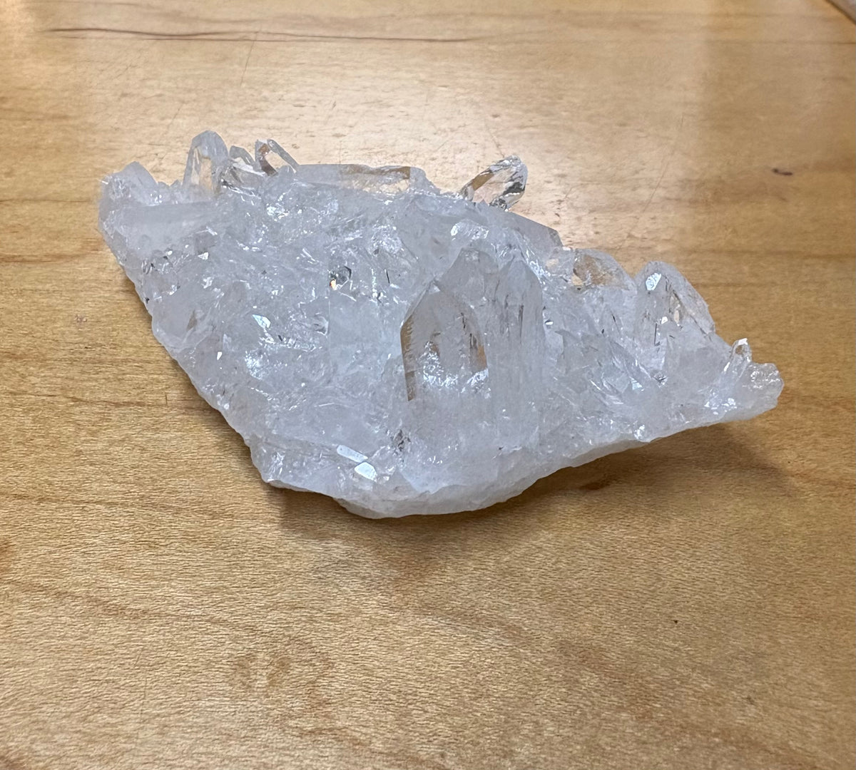 A raw, translucent clear quartz crystal cluster with visible natural points and rough edges, resting on a light brown wooden surface.