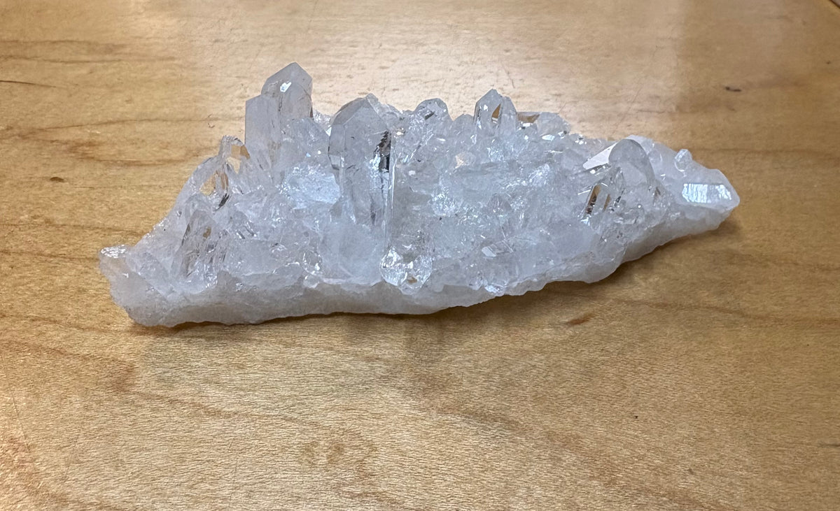 A raw white crystal cluster with multiple pointed quartz-like tips resting on a light brown wooden surface.