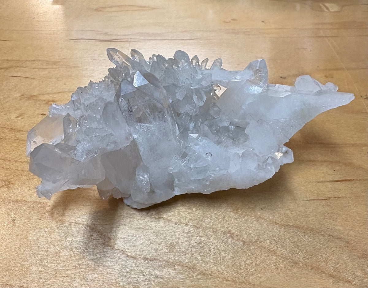 A natural clear quartz crystal cluster with multiple sharp, faceted points resting on a light-colored wooden surface.