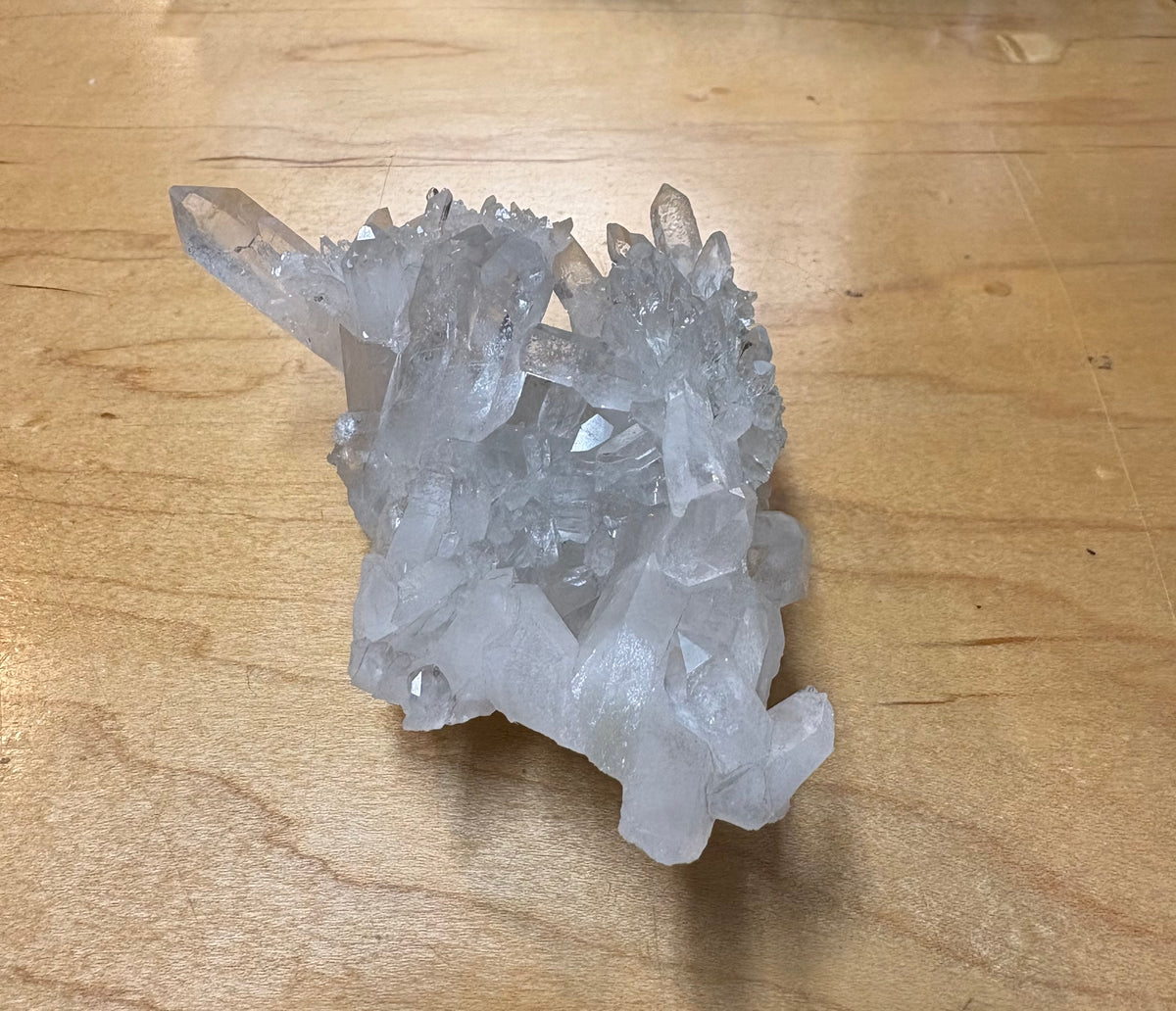 A natural clear quartz crystal cluster with multiple pointed crystal points resting on a light brown wooden surface.