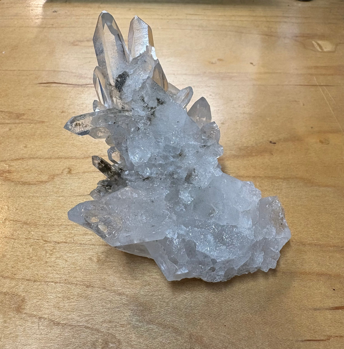 A natural clear quartz crystal cluster with multiple pointed tips and a rough, unpolished base, resting on a light brown wooden surface.