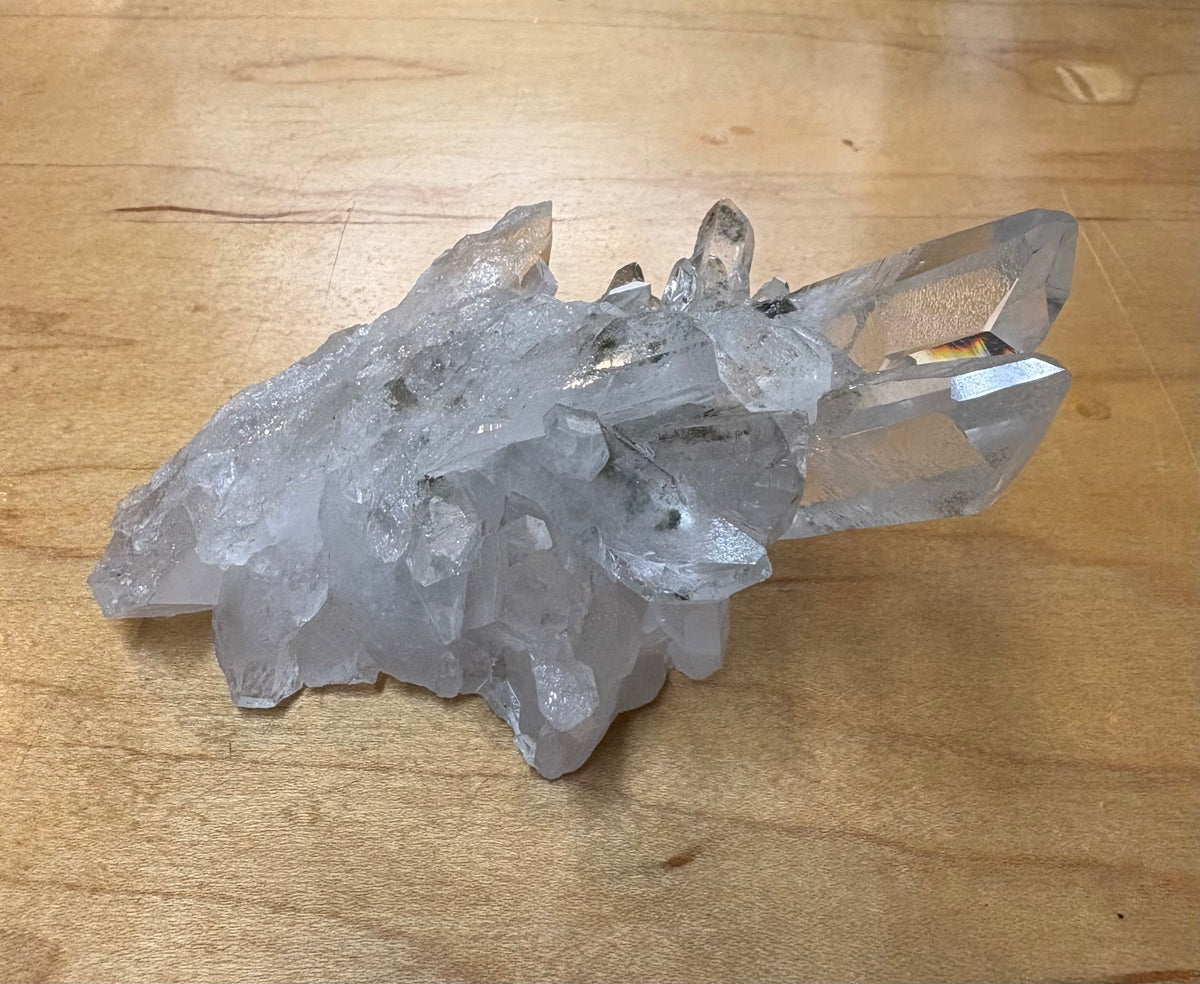 A natural clear quartz crystal cluster with multiple pointed crystal points, resting on a light brown wooden surface.