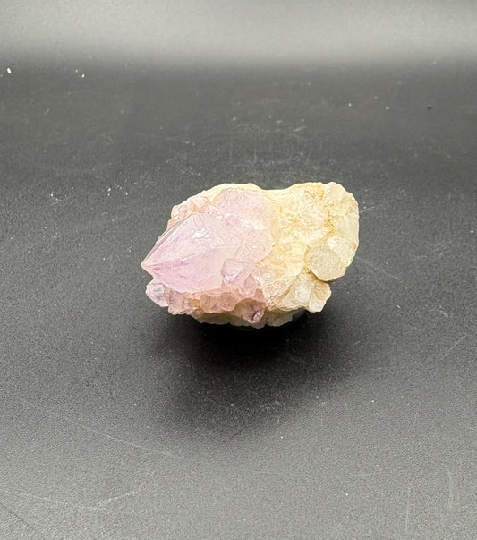 A raw mineral specimen featuring a pink Spirit Crystal point emerging from a pale cream-colored matrix, displayed on a dark gray background.