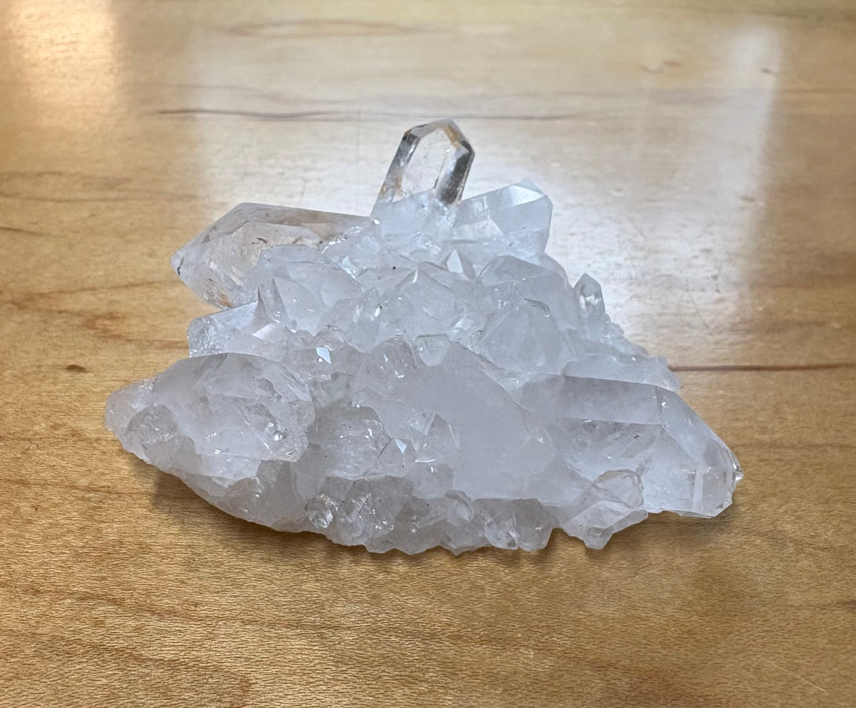 A cluster of clear quartz crystal points with a polished top segment, resting on a light brown wooden surface.