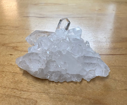 A cluster of clear quartz crystal points with a polished top segment, resting on a light brown wooden surface.