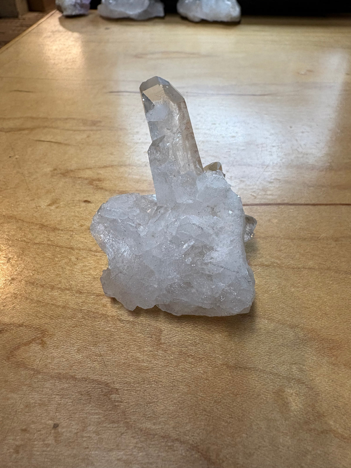A raw clear quartz crystal cluster with a tall central point resting on a light-colored wooden tabletop.