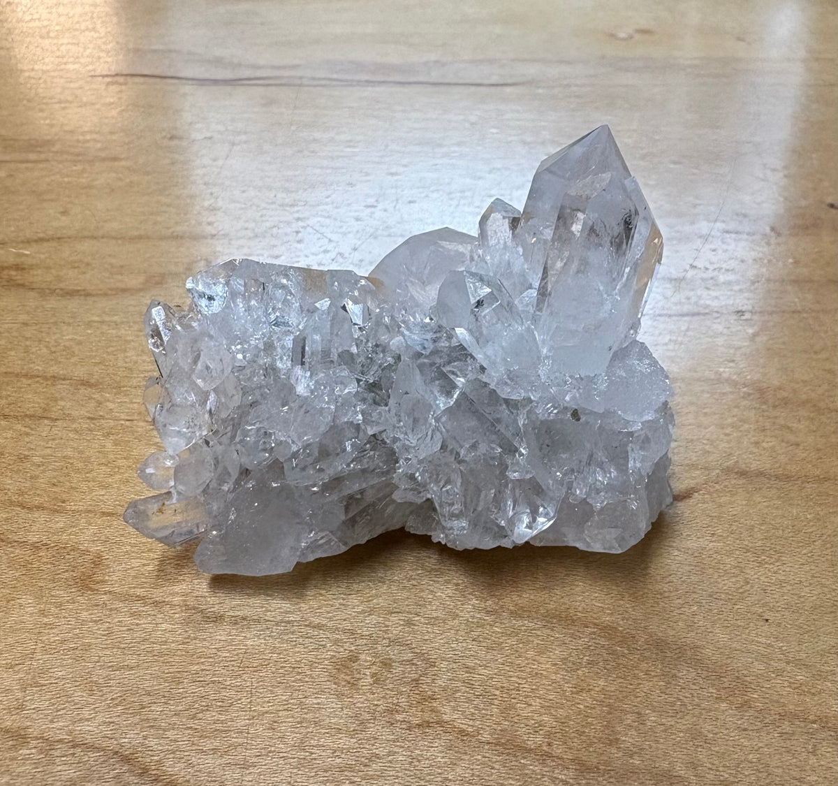 A raw crystal cluster, likely quartz, with clear faceted points resting on a light brown wooden surface.