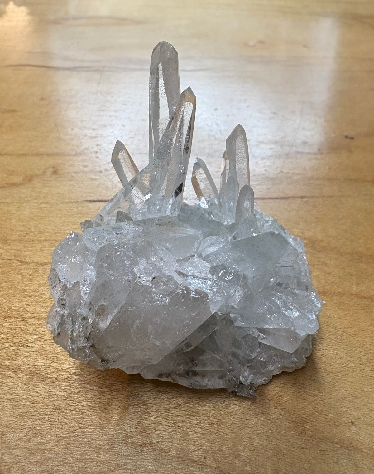 A natural clear quartz crystal cluster with multiple pointed crystal points rising from a rough, translucent base, photographed on a light wood surface.