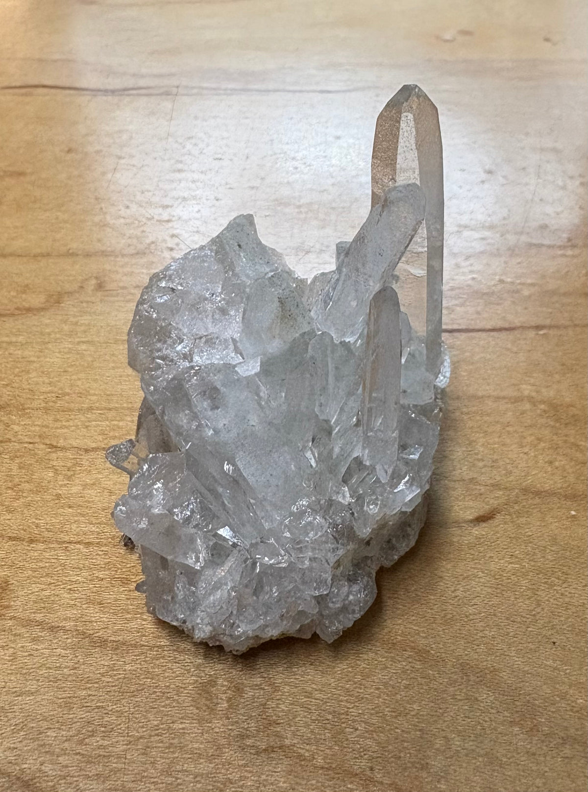 A raw clear quartz crystal cluster with a prominent pointed crystal point, resting on a light brown wooden surface.