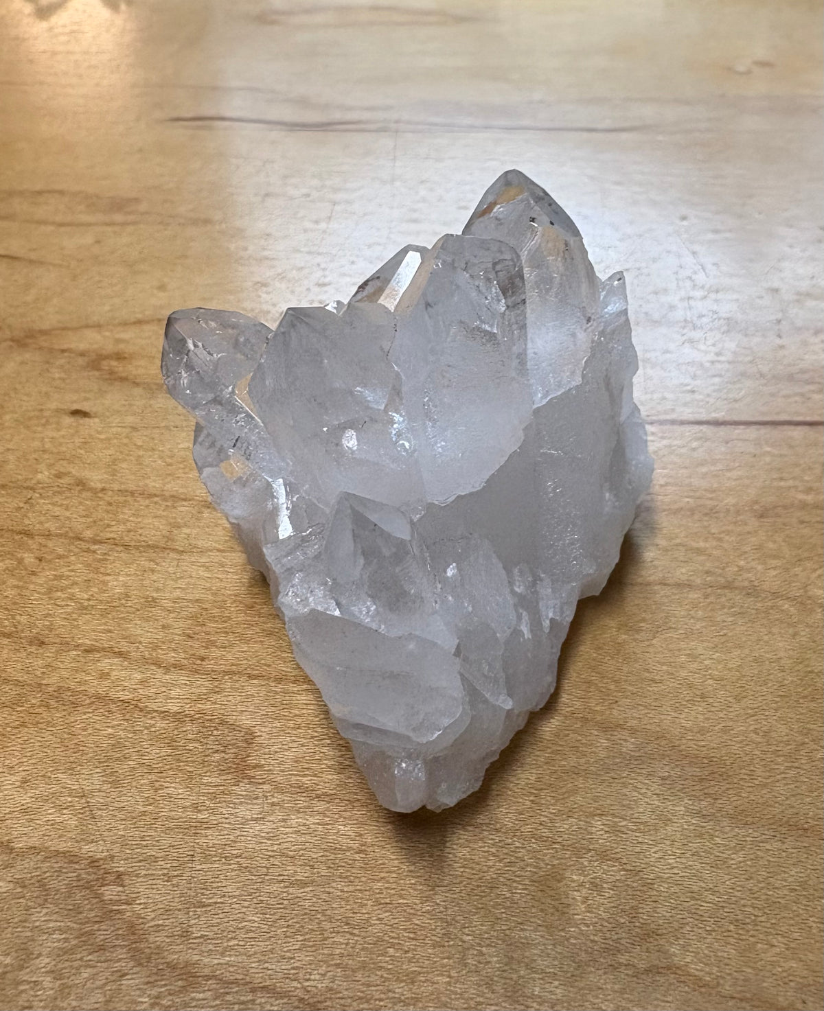 A raw, unpolished crystal cluster with multiple sharp points, showing a translucent milky white color, resting on a light brown wooden surface.
