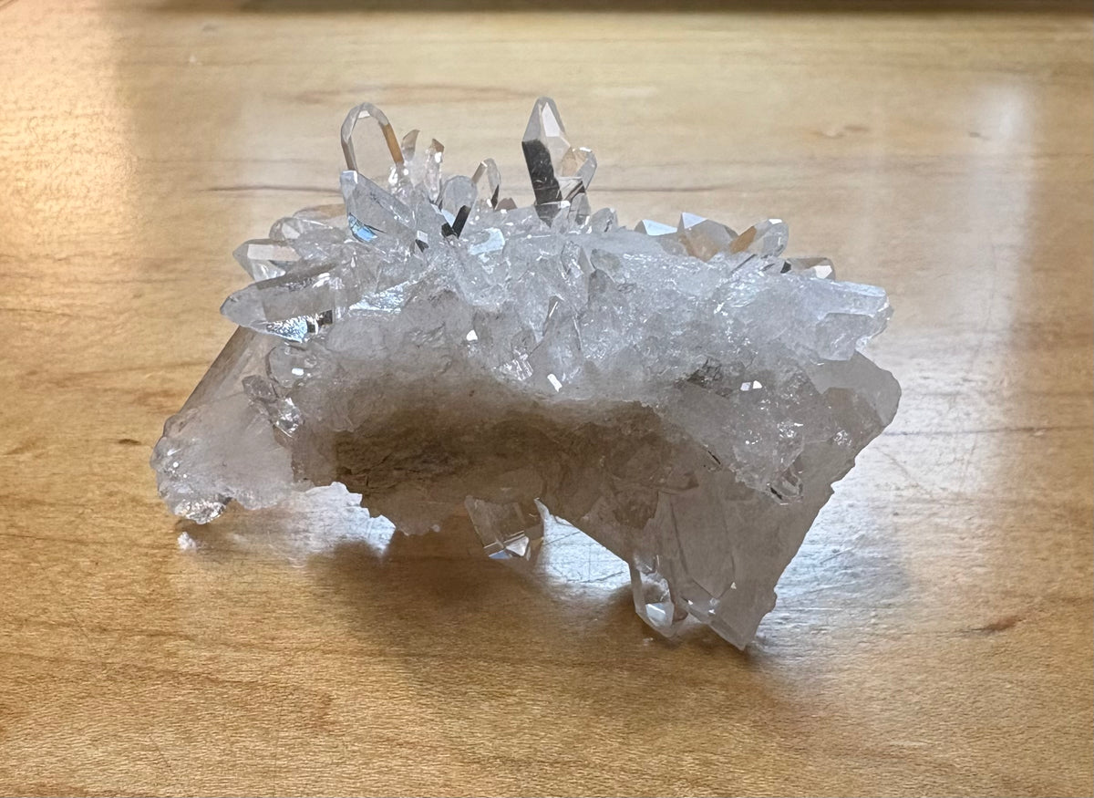 A natural clear quartz crystal cluster with multiple pointed tips, resting on a light brown wooden surface.