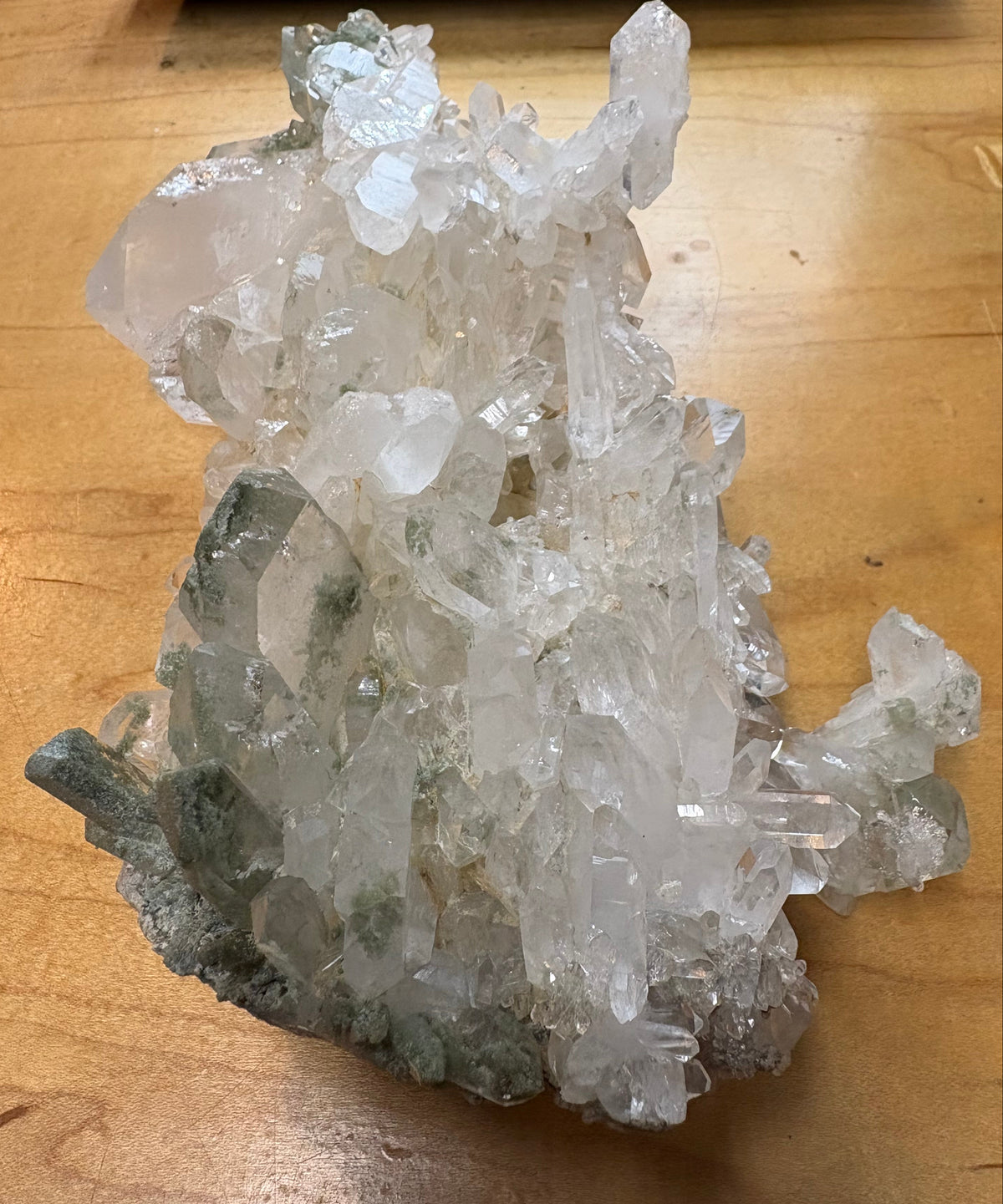 A natural crystal cluster with clear and translucent quartz-like points and some gray-green matrix, photographed on a wooden tabletop.