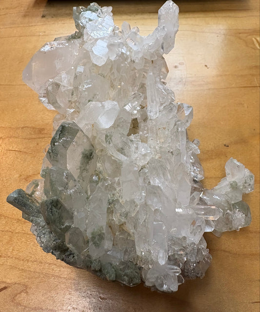 A natural crystal cluster with clear and translucent quartz-like points and some gray-green matrix, photographed on a wooden tabletop.