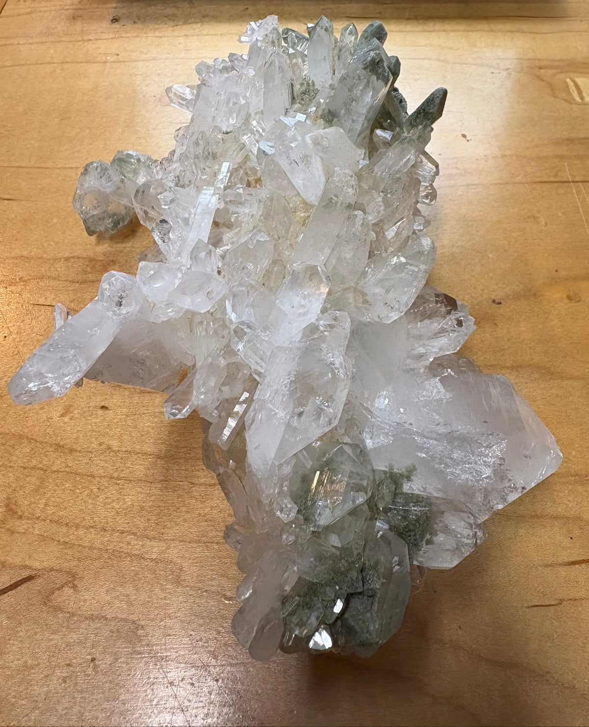 A large natural quartz crystal cluster with clear, faceted points and subtle gray-green inclusions, displayed on a wooden tabletop.