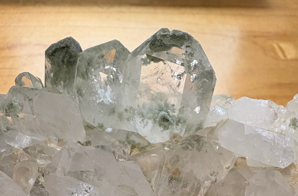 A close-up of a clear quartz crystal cluster with prominent greenish impurities inside the larger crystals, resting on a light wooden surface.