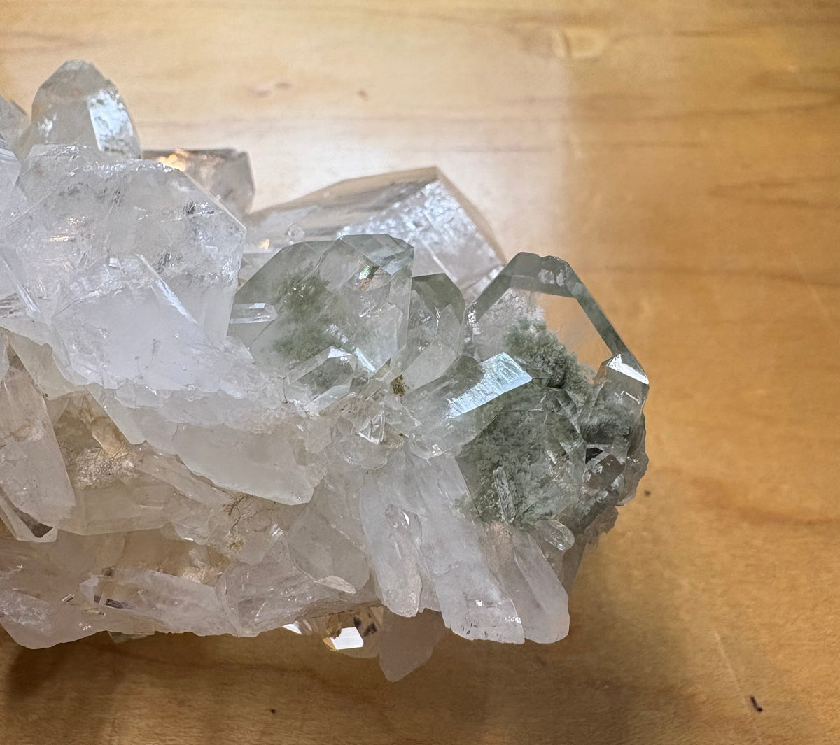 A natural clear quartz crystal cluster with some greenish mineral inclusions, resting on a light brown wooden surface.